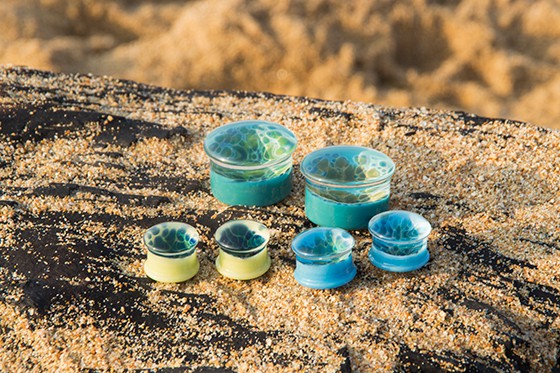 Glass or plastic plugs of various sizes with a blue ocean/wave design, sitting on a sandy wooden bench on the Sunshine Coast
