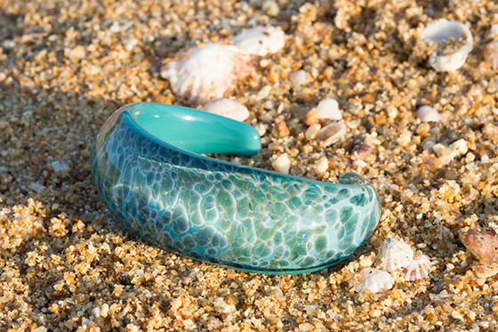 Turquoise bracelet with a wave/ocean design sitting on a sandy Sunshine Coast beach surrounded by shells.