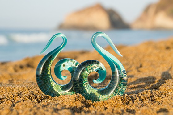 A pair or blue and green ocean/wave design spiral ear stretchers/tapers, sitting on the sand on the Sunshine Coast